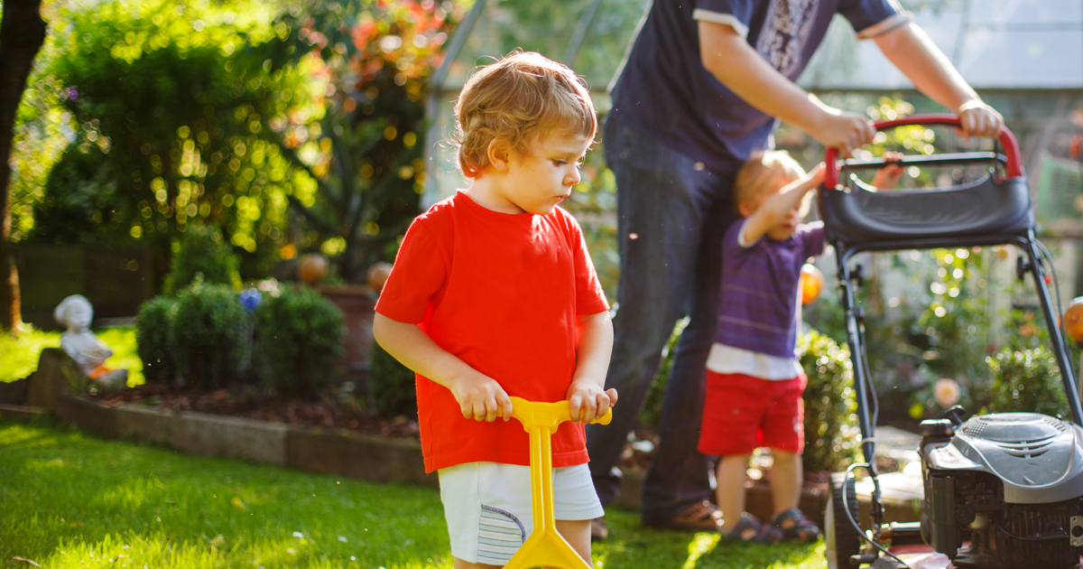 Photo of a child playing with toy near adult mowing the lawn with a lawnmower