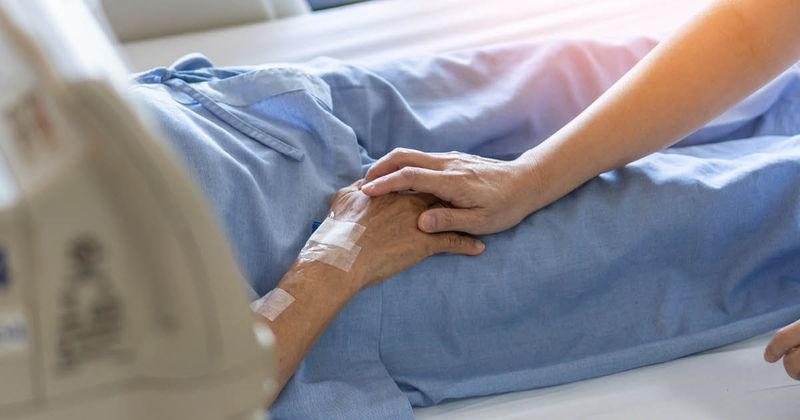 Caregiver holding older patient's hand while they are in a hospital bed.