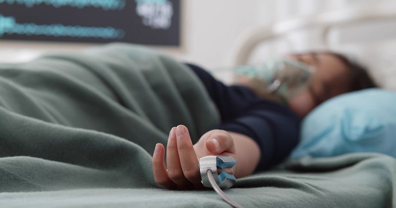 Teen Asian girl wearing an oxygen mask and an oximeter while sleeping in a hospital bed.