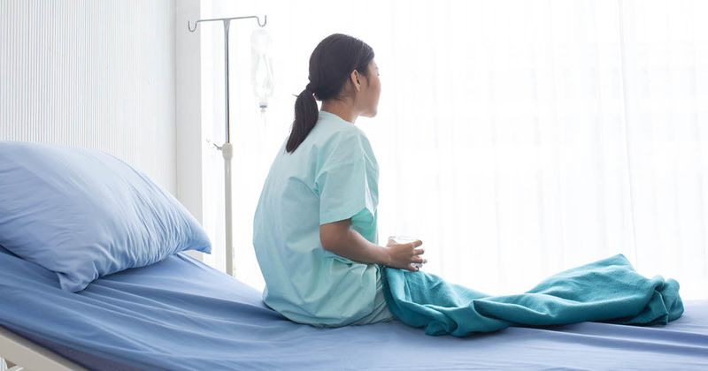 Asian female patient sat on a hospital bed holding water and looking out the window with sad emotions.