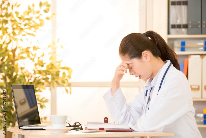 Female doctor sitting at desk rubbing her eyes with computer screen at left and glasses in foreground